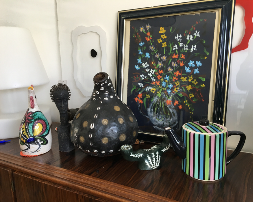 Une collection d'objets décoratifs éclectiques sur un buffet en bois, dont une figurine de coq colorée d'époque, une sculpture africaine noire, une théière à pois, une tasse rayée vintage