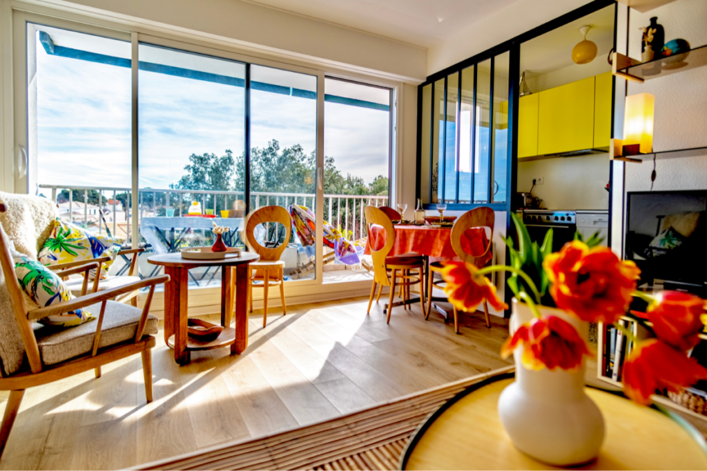 Salon lumineux et moderne avec de grandes fenêtres donnant sur une piscine dans Location saisonnière à Pornic, comprenant un coin repas coloré et une cuisine aux armoires jaunes.