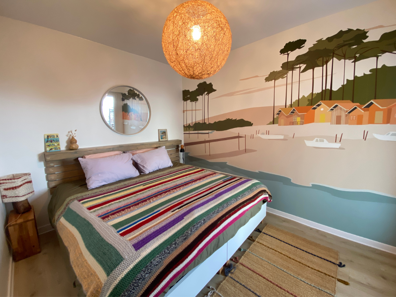 Cette charmante chambre située dans un emplacement saisonnier à Pornic comprend un grand lit drapé d'une couverture rayée, avec une fresque murale de maisons et d'arbres ornant le mur. Un miroir rond et un luminaire suspendu sphérique ajoutent à l’ambiance chaleureuse.