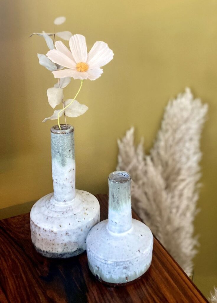 Deux vases en céramique blanche sont disposés sur une surface en bois. L'un des vases en grès contient une seule fleur blanche et quelques feuilles. De l'herbe de la pampa sèche est visible à l'arrière-plan sur un mur vert clair.
