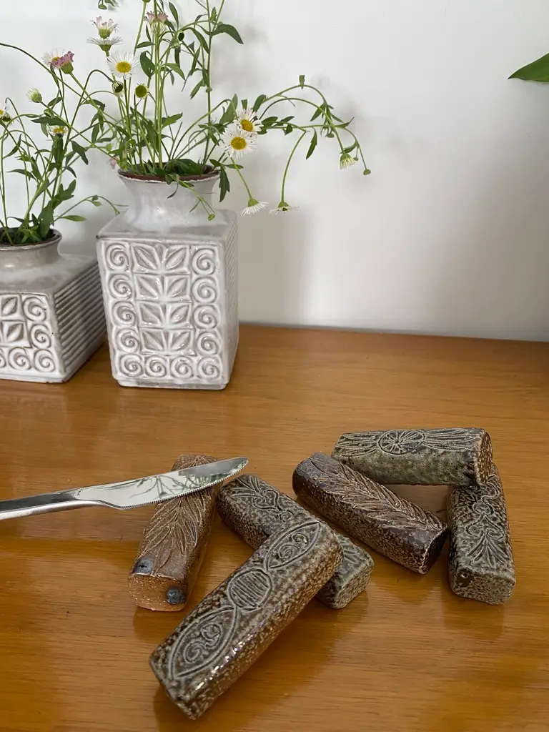 Des tampons en bois sculptés à la main reposent sur une surface en bois à côté d'un couteau à beurre, avec des porte-couteaux en grès soutenant le décor et des poteries décoratives tenant des fleurs en arrière-plan.